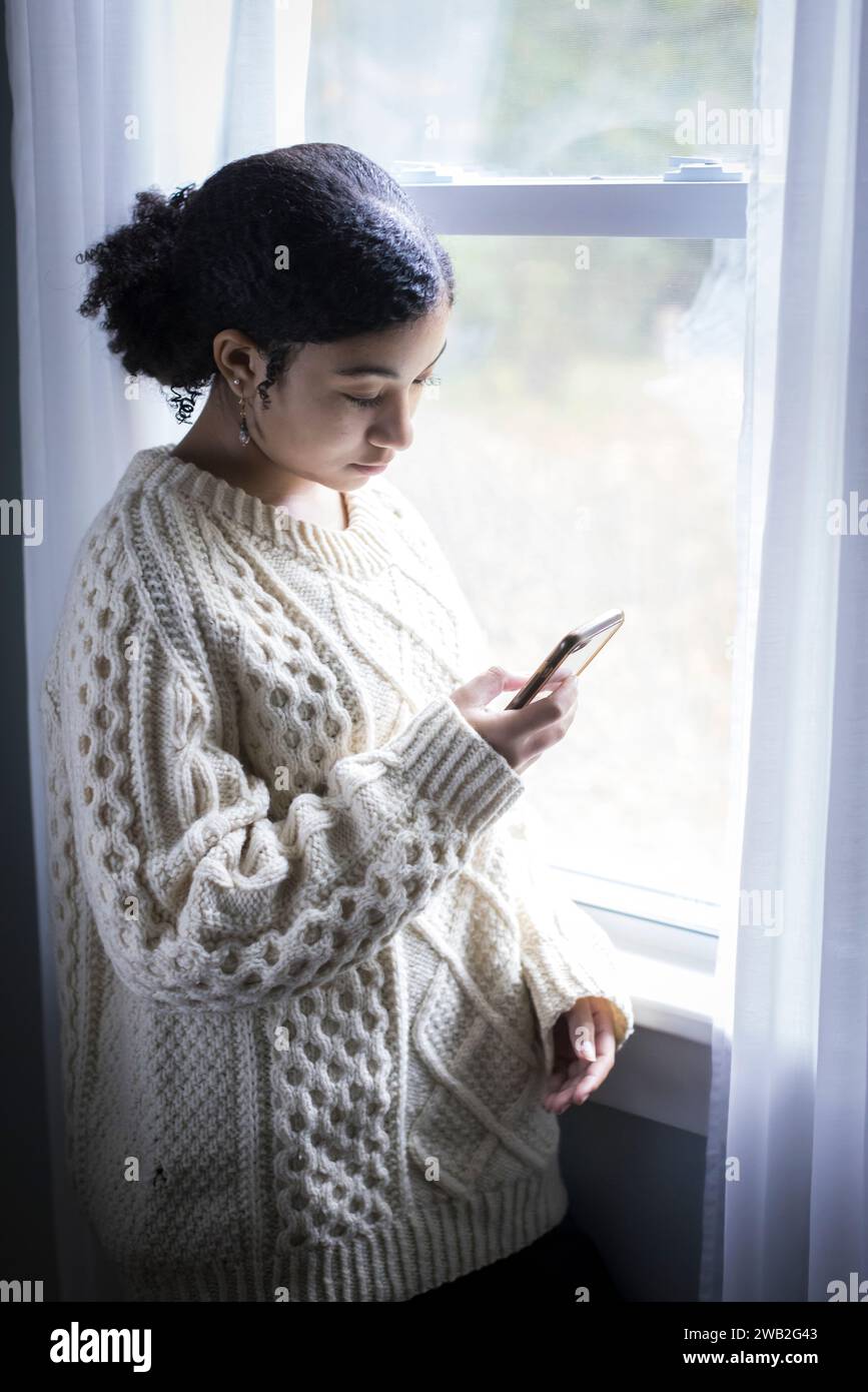 La ragazza adolescente birazziale guarda il suo cellulare dalla finestra Foto Stock