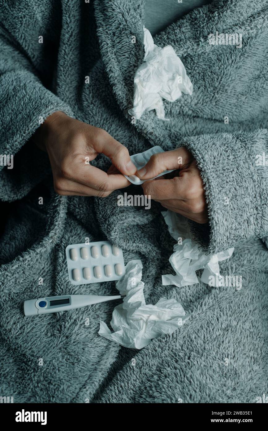 un uomo caucasico malato a casa, indossando un morbido abito grigio casa, circondato da tessuti usati, sta per prendere una pillola Foto Stock