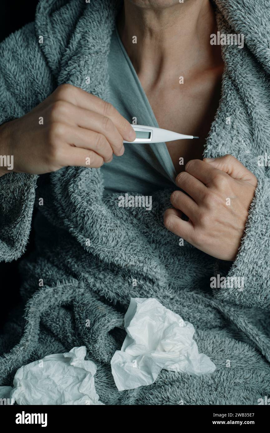 un uomo malato a casa, che indossa un soffice abito da casa grigio, sta per misurare la sua temperatura con un termometro digitale, circondato da tessuti usati Foto Stock