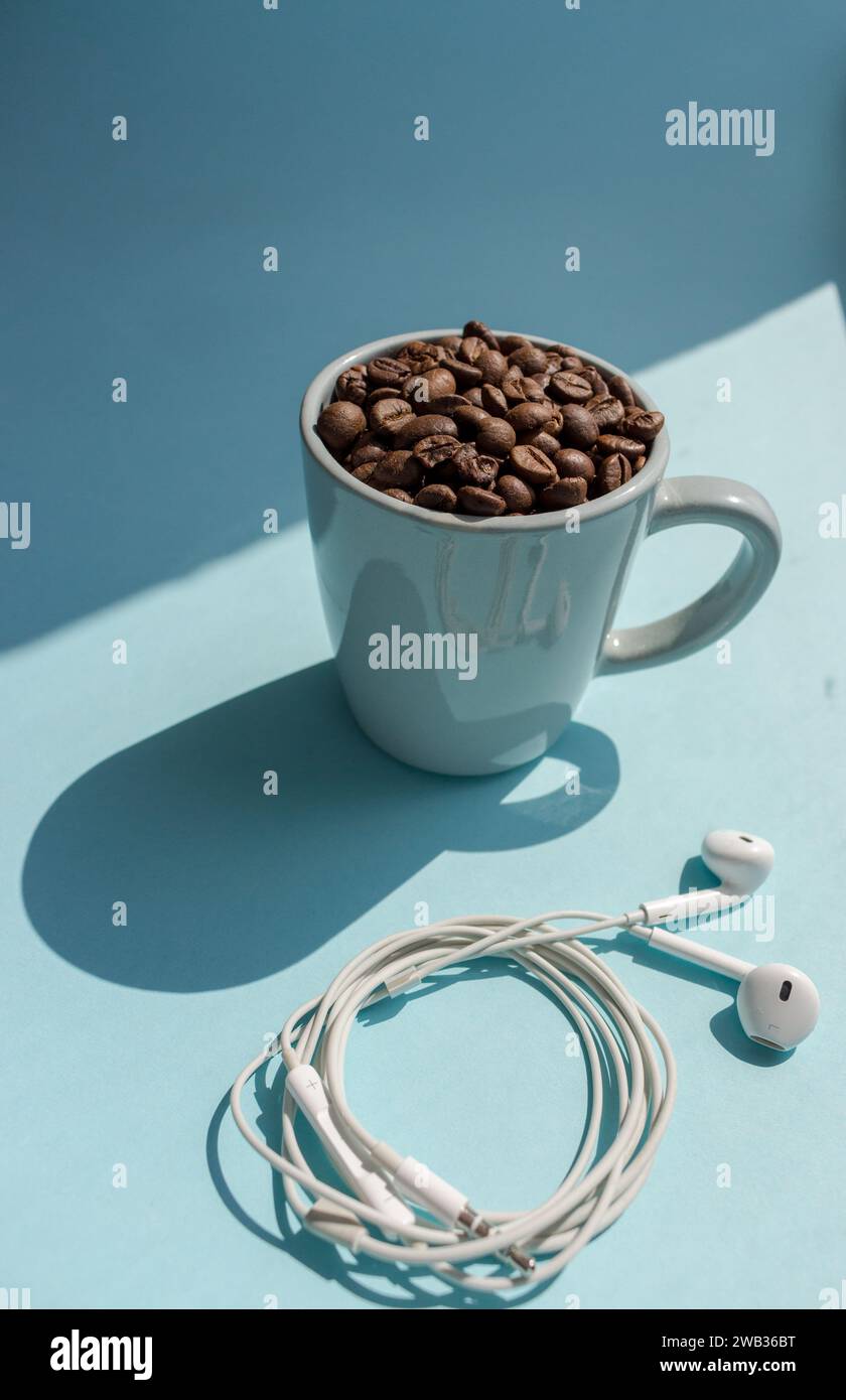 Chicchi di caffè neri in tazza blu e cuffie bianche su sfondo blu con vista dall'alto con ombre solari. Drink mattutino. Caffè e musica. Foto Stock