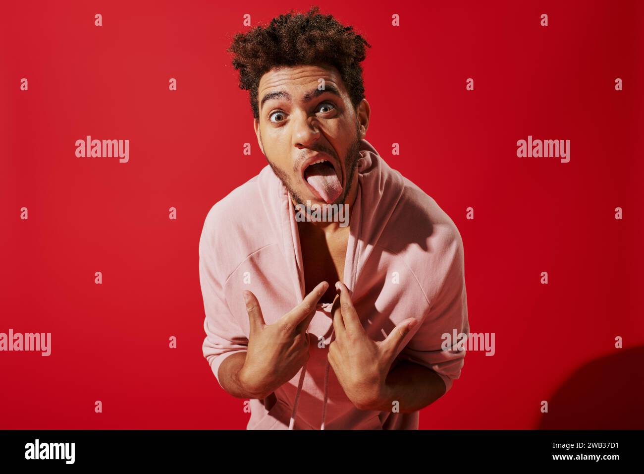 simpatico afroamericano che tira la felpa con cappuccio rosa e si staglia la lingua mentre si trova su sfondo rosso Foto Stock