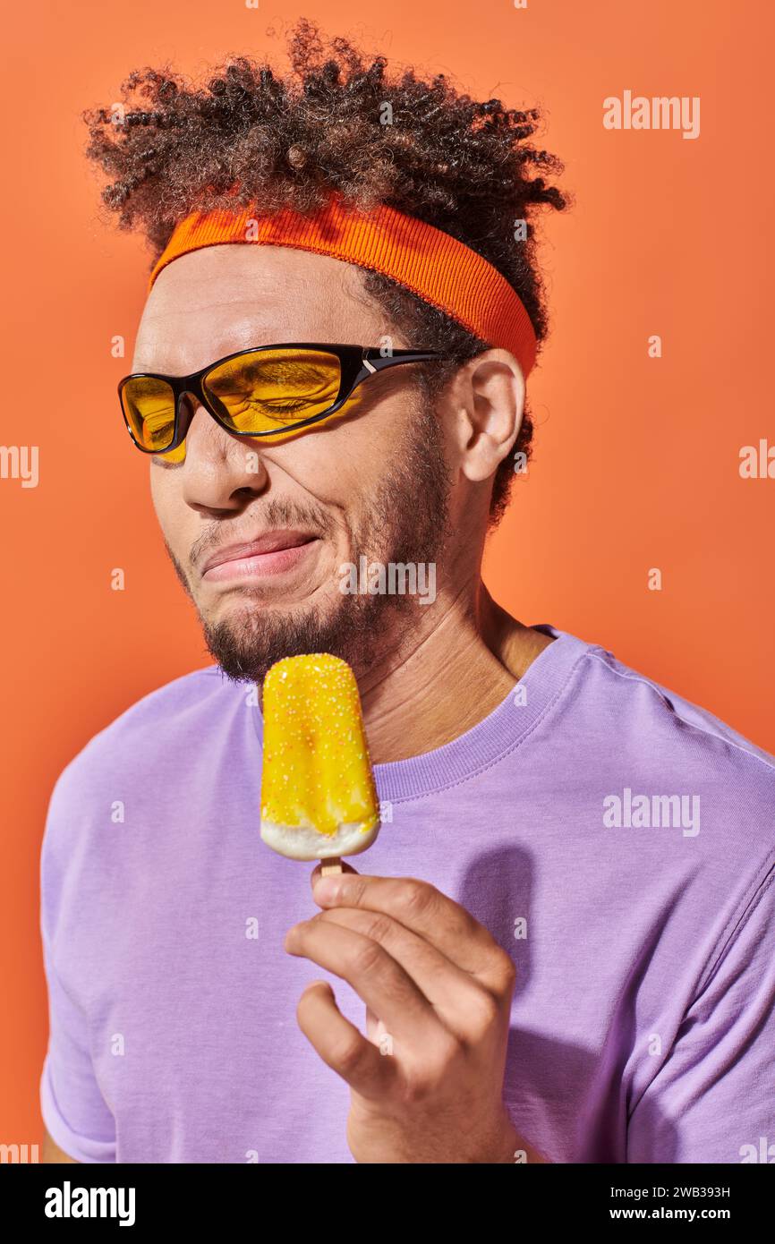 uomo afro-americano con occhiali da sole e cerchietto che grigia con gelato acido su sfondo arancione Foto Stock