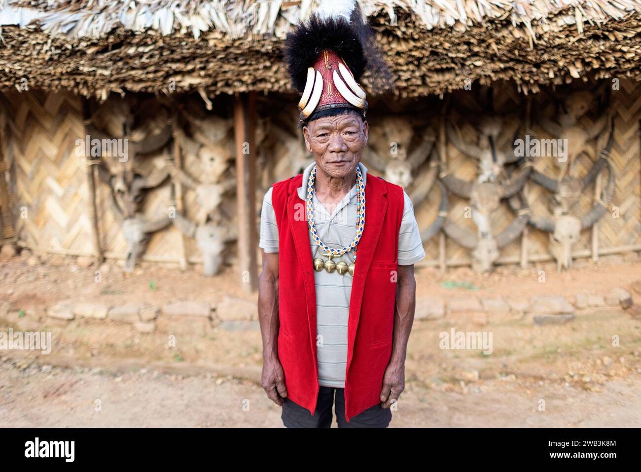 Anziano della tribù Konyak in abiti cerimoniali tradizionali, in piedi davanti a una casa in legno konyak, decorata con teschi di toro, Nagaland, India Foto Stock