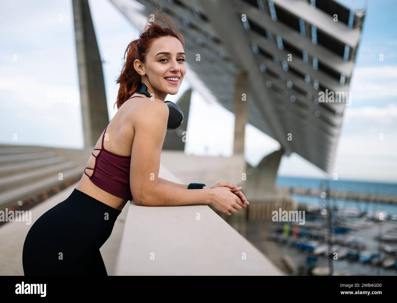 Giovane donna in forma sorridente che indossa un abbigliamento sportivo e che indossa cuffie esterne che guardano la fotocamera. Atletica femminile che ascolta musica che respira durante un Foto Stock