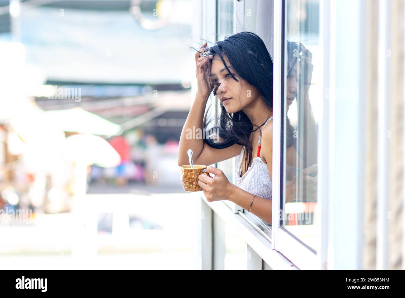 Una donna con un vestito estivo tiene una tazza in una finestra aperta di un edificio residenziale mentre il vento soffia attraverso i capelli Foto Stock