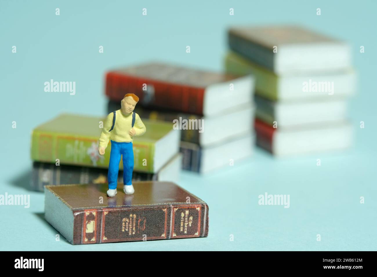 Fotografia in miniatura di giocattoli per persone piccole. Un ragazzo con uno zaino scolastico in piedi sopra la scala del libro. Isolato su sfondo blu. Foto immagine Foto Stock