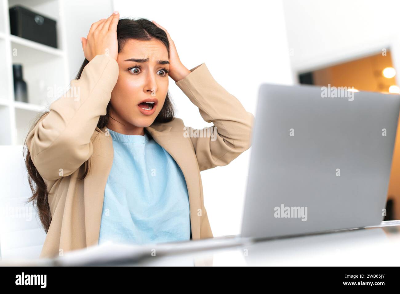Scioccata, perplessa, donna d'affari indiana o araba, seduta a una scrivania in ufficio, sguardi confusi sullo schermo del notebook, afferra la testa con le mani, è perplessa Foto Stock
