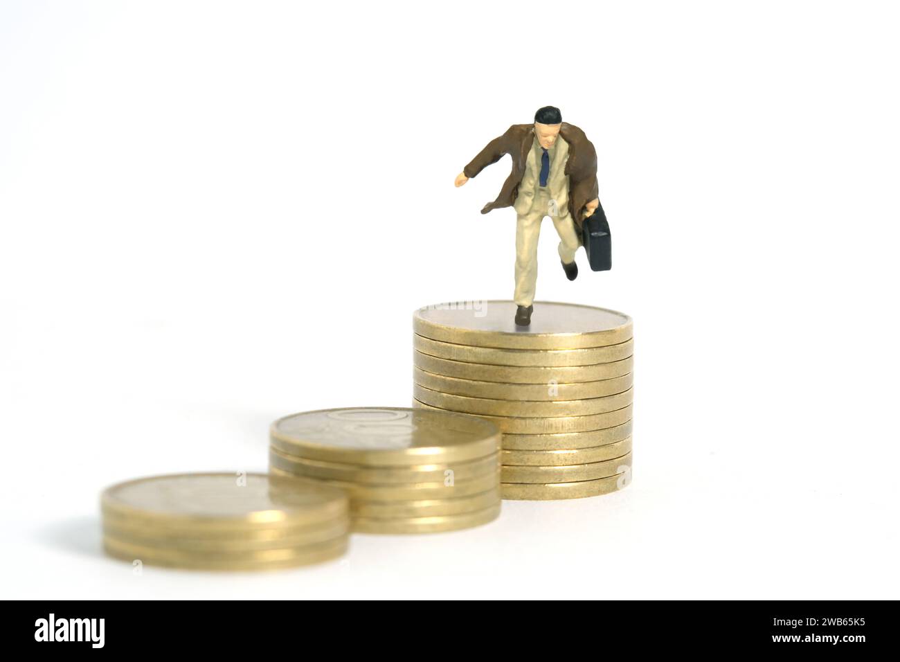 Fotografia di figure giocattolo in miniatura. Un uomo d'affari che indossa un cappotto che salta sopra la pila di monete dorate. Isolato su sfondo bianco. Foto immagine Foto Stock
