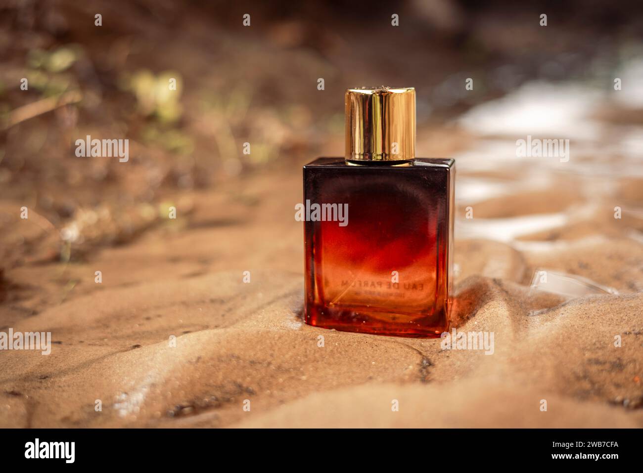 Una bottiglia di profumo di lusso color arancio scuro dorato conservata nella sabbia bagnata. Foto Stock