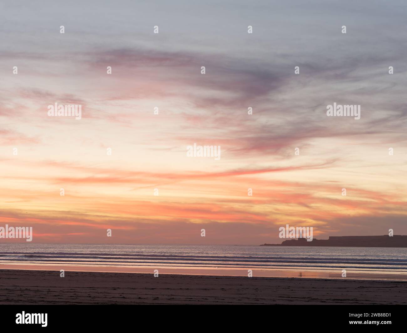 Nuvole spettacolari al tramonto, mentre le onde rotolano verso la spiaggia sabbiosa con un'isola alle spalle della città di Essaouira, in Marocco. 8 gennaio 2024 Foto Stock