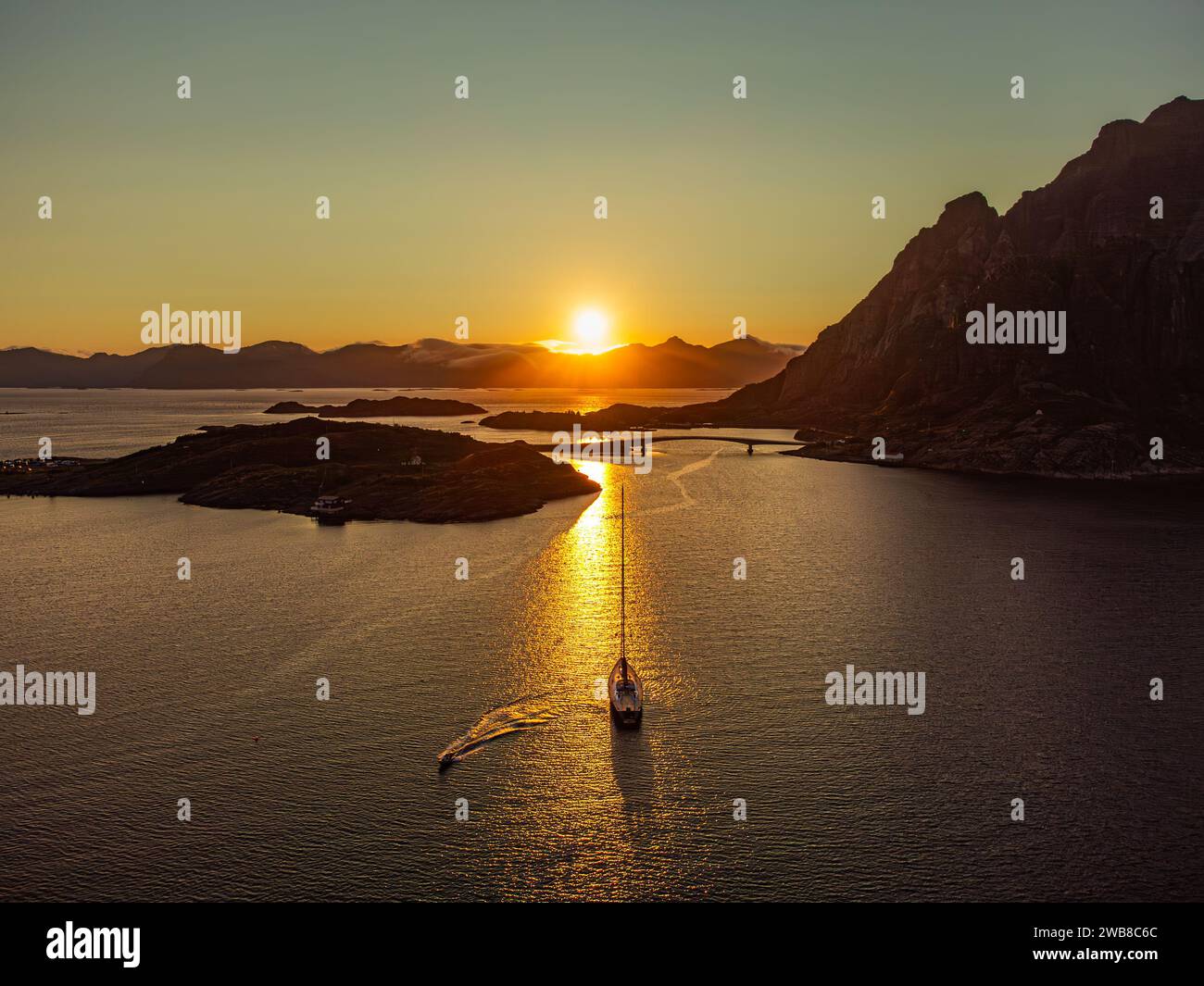 Una barca a vela sotto il sole di mezzanotte, dietro una grande collina a Lofoten, vicino a Henningsvaer, in lontananza alte montagne, ponte che collega Henningsvaer. Foto Stock