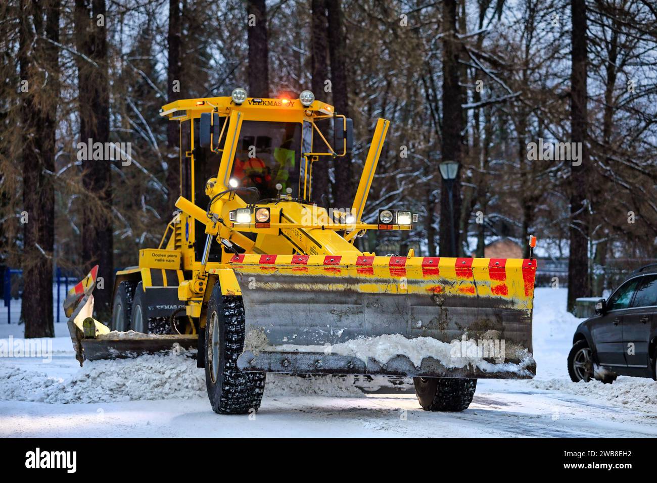 Motorgrader giallo Veekmas FG 2327 S che rimuove la neve dal parcheggio con lama regolabile dopo le nevicate invernali. Salo, Finlandia. 27 dicembre 2023. Foto Stock