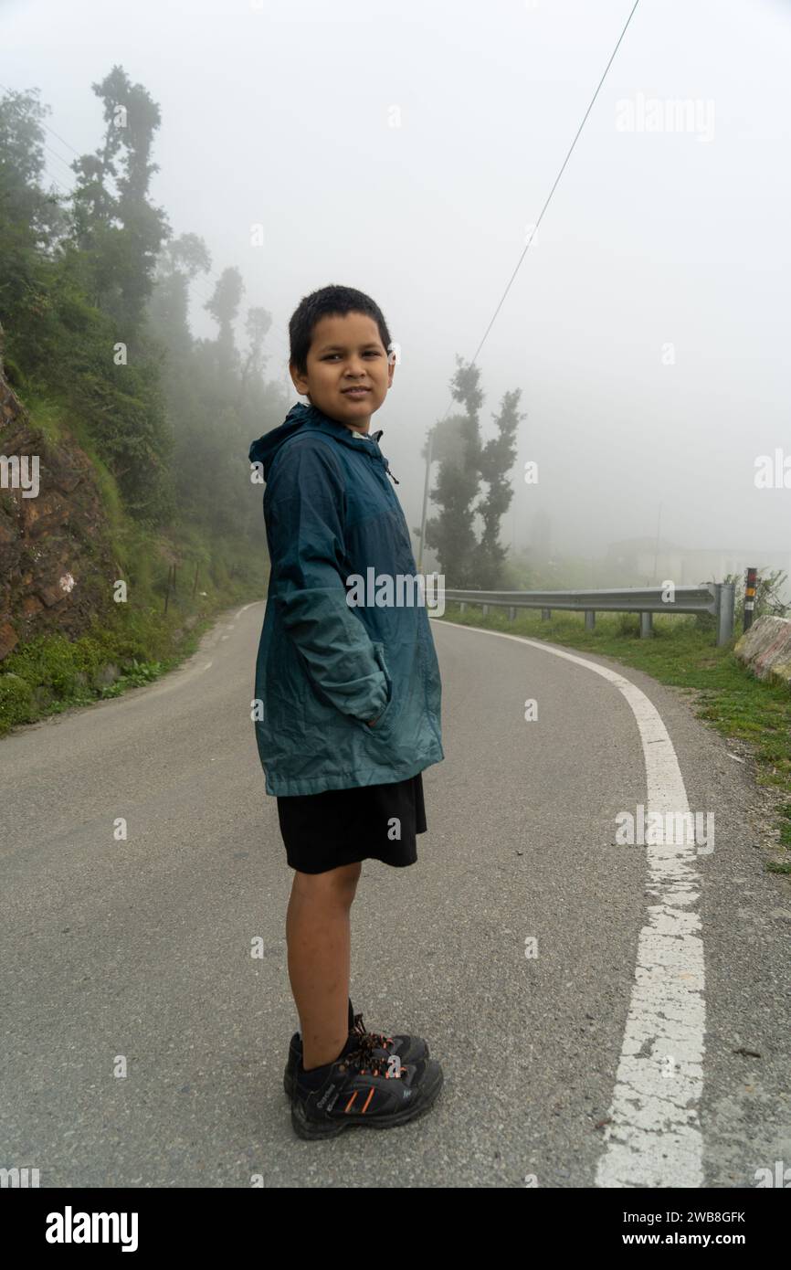 Nei pittoreschi paesaggi di Uttarakhand, un giovane ragazzo si trova accanto alla strada, il suo viaggio si svolge sullo sfondo di una bellezza paesaggistica. Foto Stock