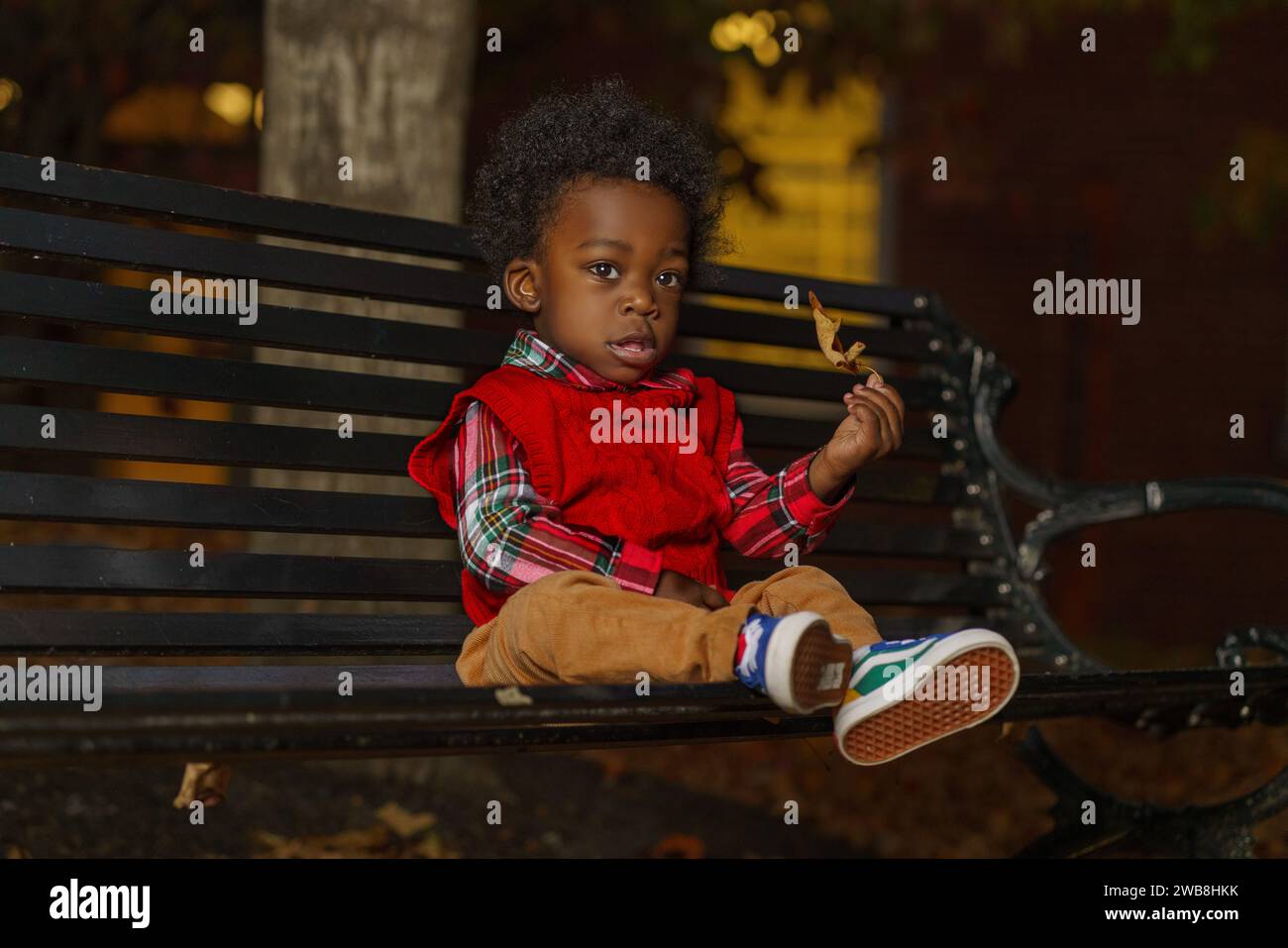 Un bambino africano è seduto sopra una panchina di legno, curiosamente guardando la macchina fotografica Foto Stock