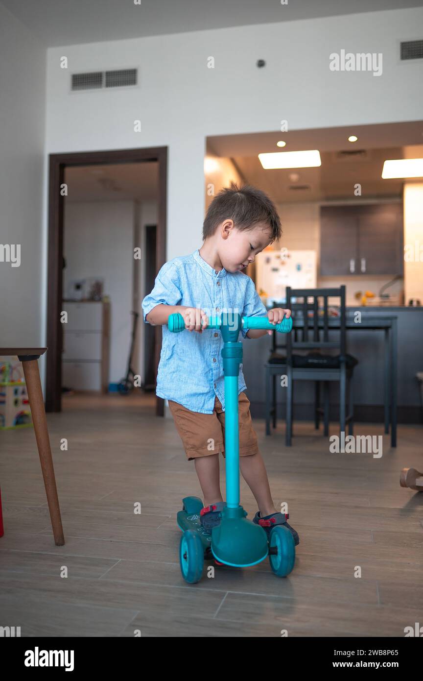 Prova la gioia pura di un bambino di due anni e mezzo, vestito con una camicia e bermuda, mentre esplora giocosamente la stanza con il suo baby push sc Foto Stock