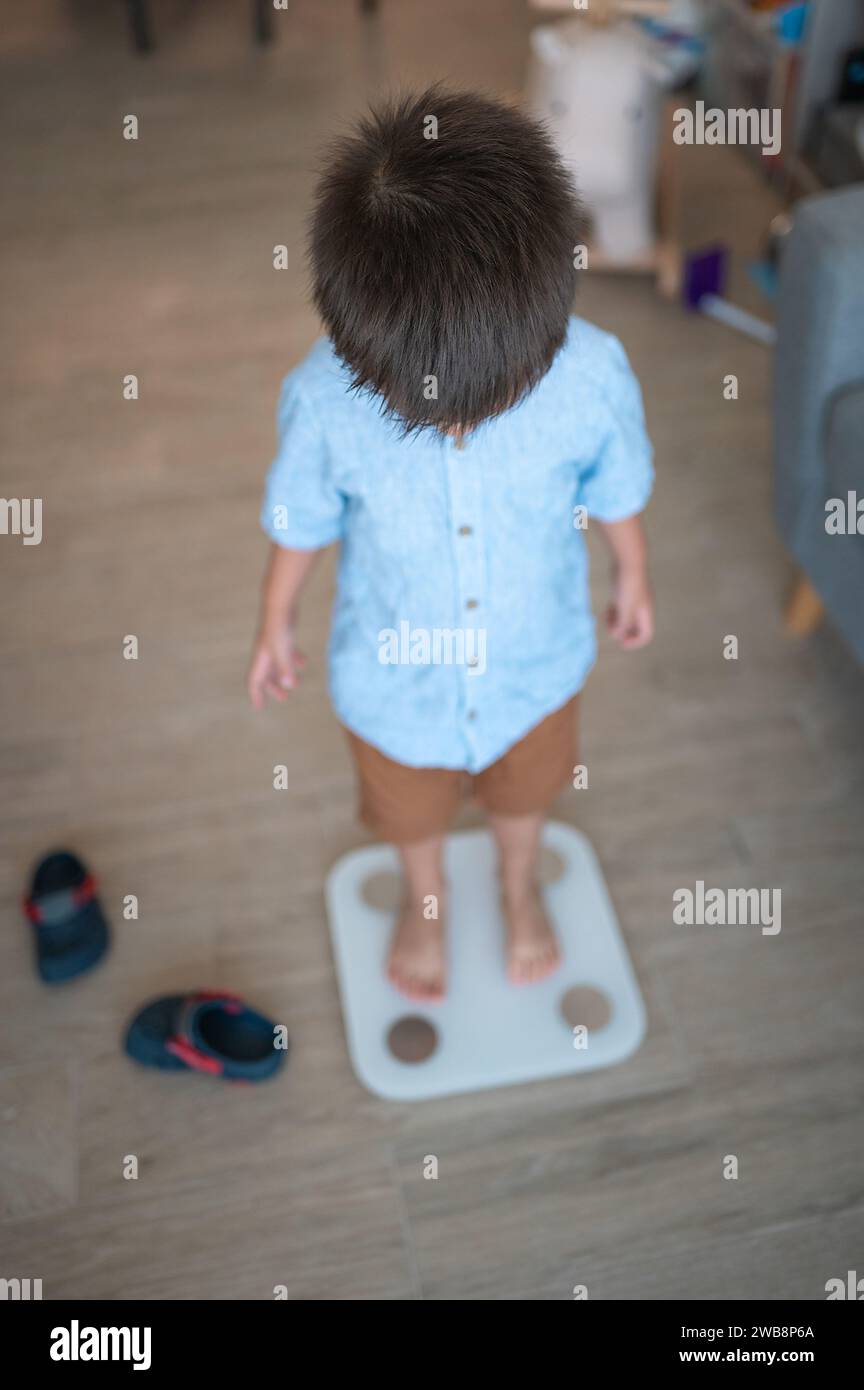 Bambino di due anni e mezzo, vestito con una camicia e bermuda, che fa un passo verso abitudini sane. In piedi scalzi su una smart scale i digitale Foto Stock