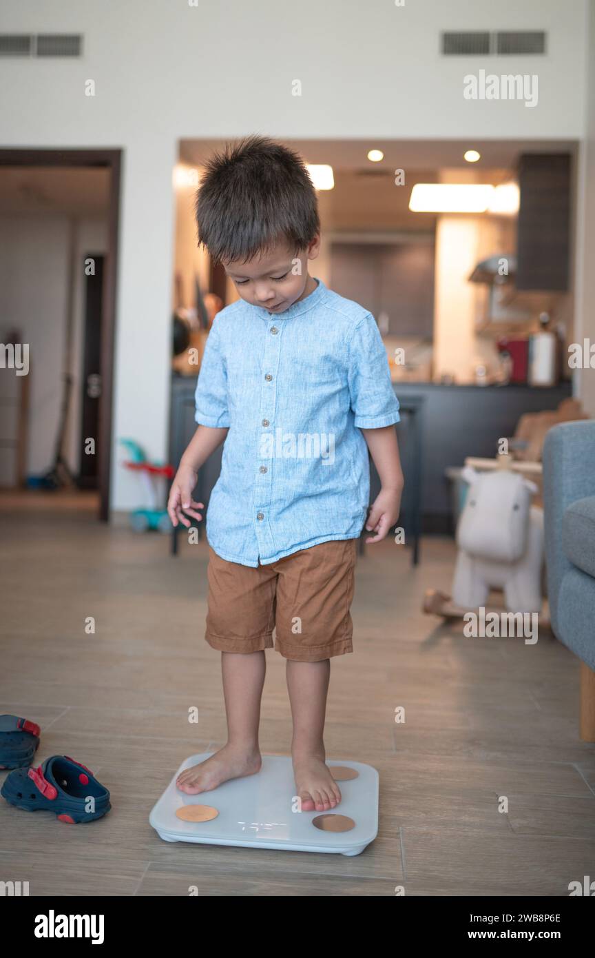 Bambino di due anni e mezzo, vestito con una camicia e bermuda, che fa un passo verso abitudini sane. In piedi scalzi su una smart scale i digitale Foto Stock