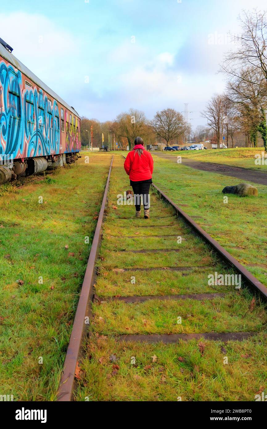 AS, Limburgo, Belgio. 17 dicembre 2023. Binari ferroviari in disuso della vecchia stazione con una donna che cammina con il suo cane, torna alla telecamera, autovetture dipinte W. Foto Stock