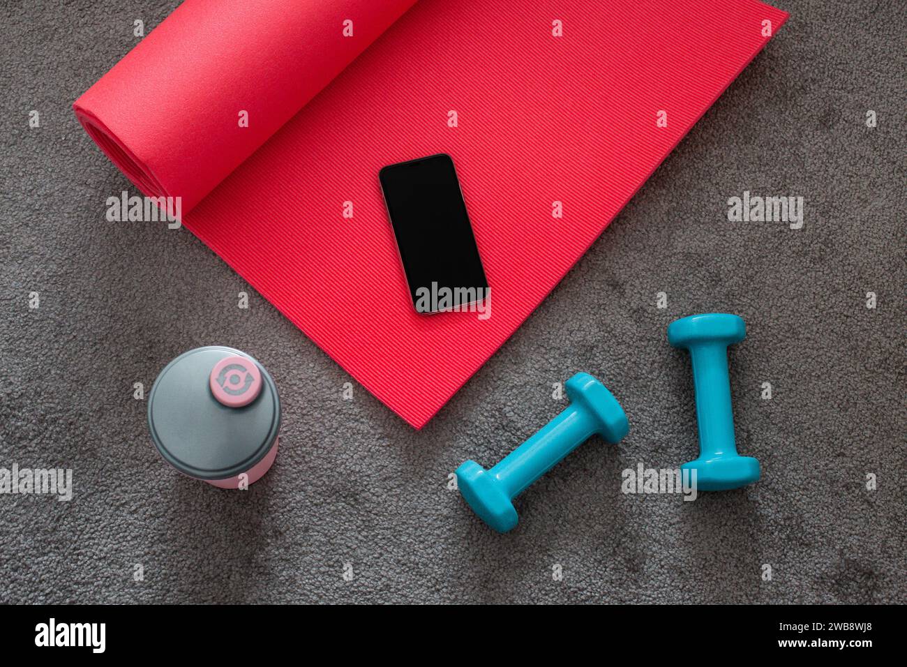 Vista dall'alto del tappetino yoga rosa, manubri blu, attrezzature fitness su tappeto grigio con telefono cellulare. Foto Stock