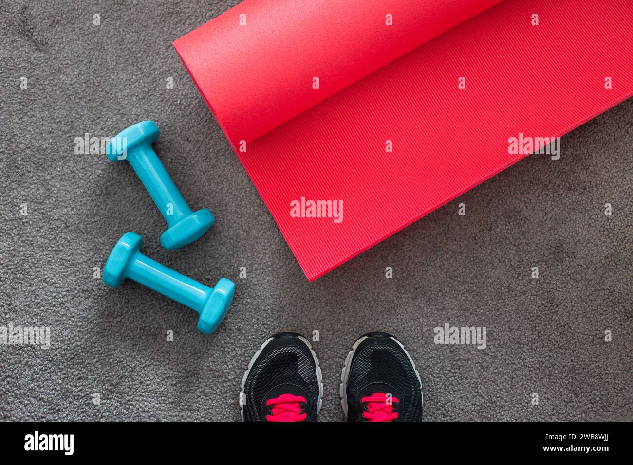 Vista dall'alto del tappetino yoga rosa, manubri blu, attrezzature fitness su tappeto grigio con scarpe. Foto Stock