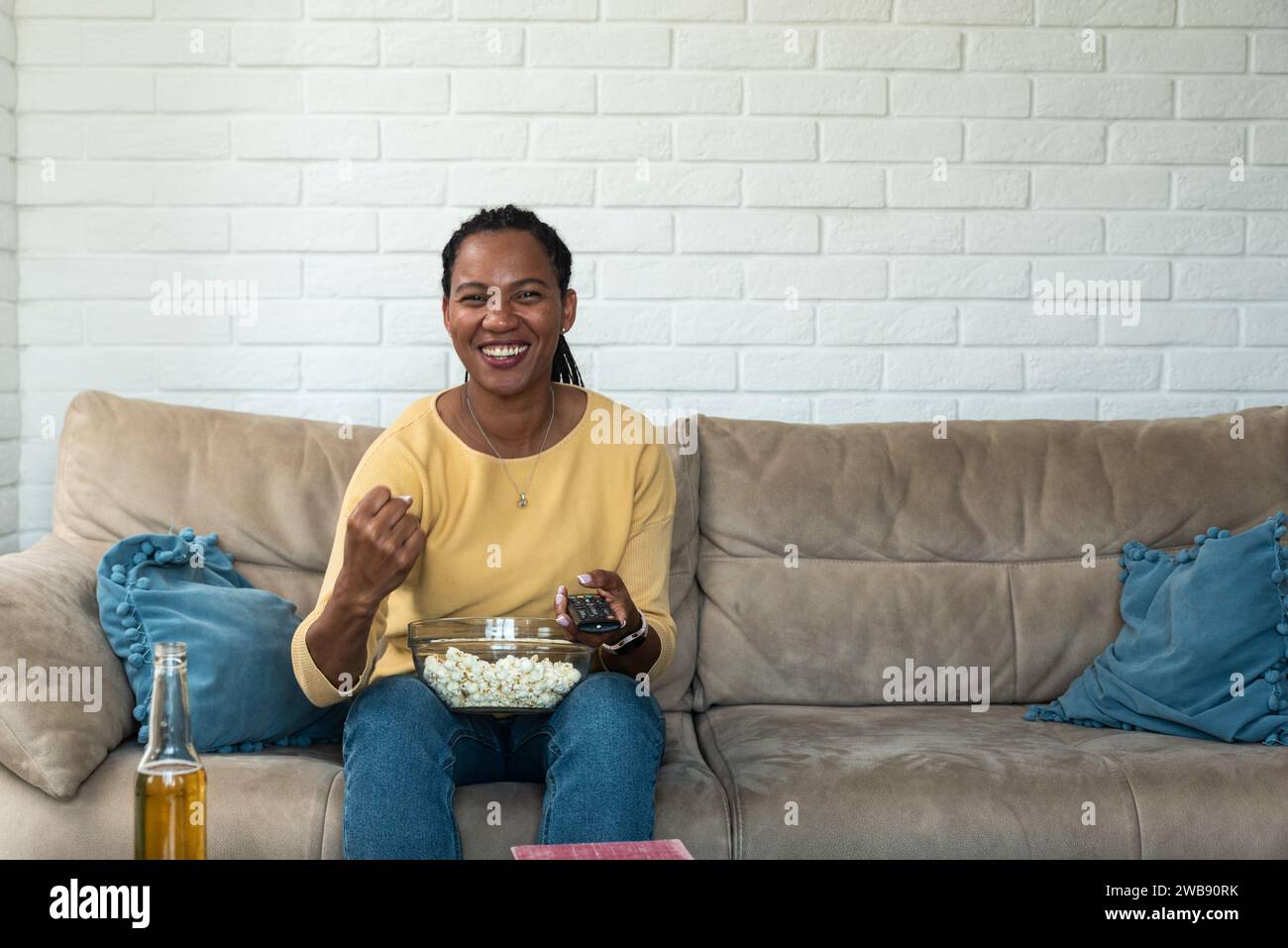 Giovane donna afroamericana seduta a casa sul divano a guardare una partita sportiva in TV, a mangiare popcorn e a fare il tifo. Madre che si gode il tempo da sola per se stessa Foto Stock