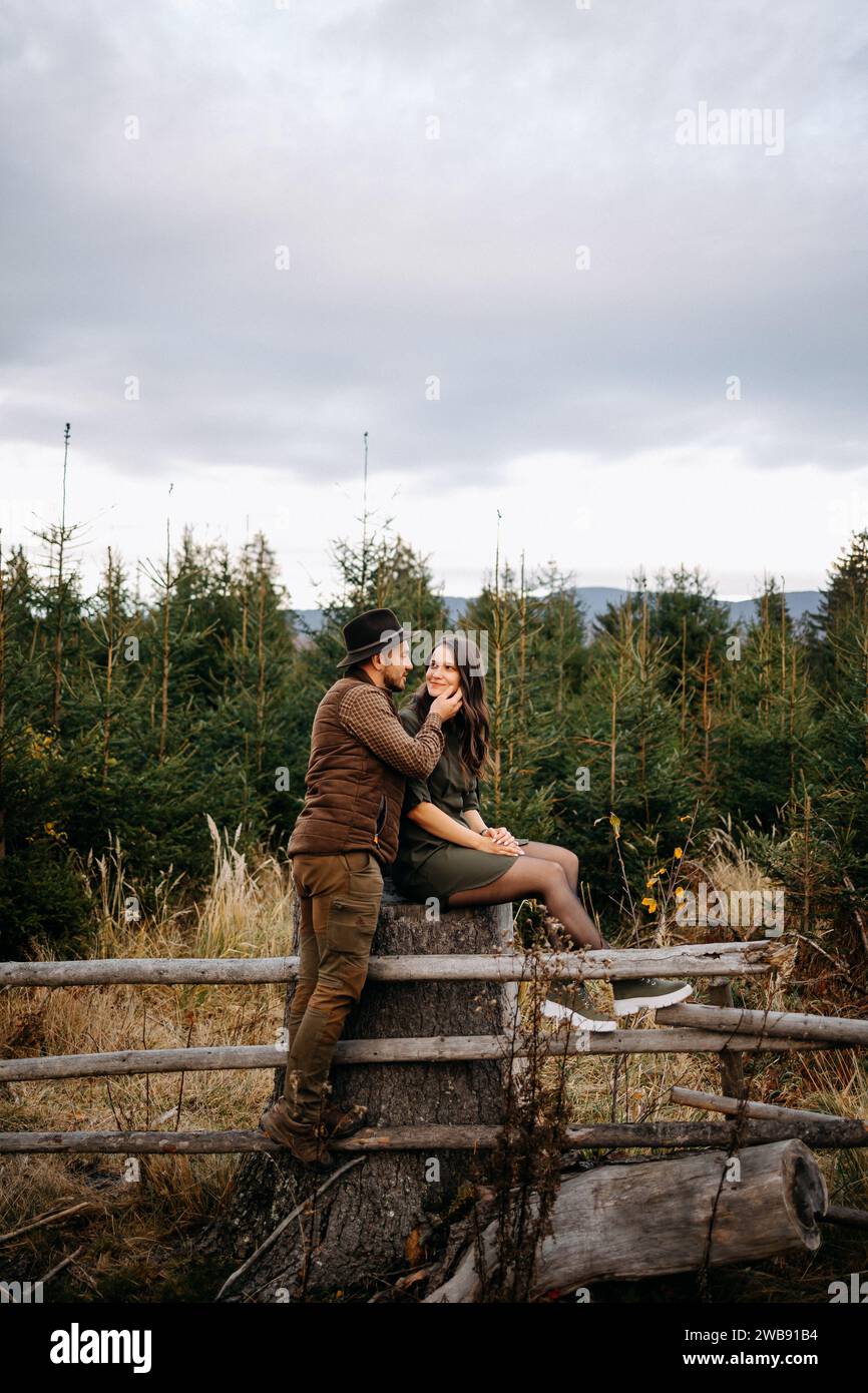 Una giovane e adorabile coppia, il cacciatore e la sua signora, si guardano e si siedono accoccolati insieme su una recinzione nella natura Foto Stock