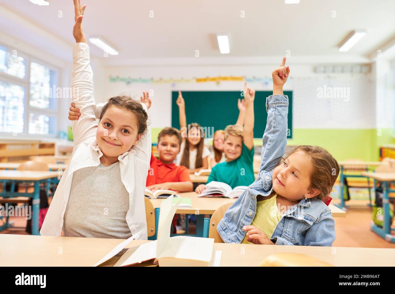 Studentessa sorridente che solleva la mano mentre si siede con un'amica durante la lezione in classe Foto Stock