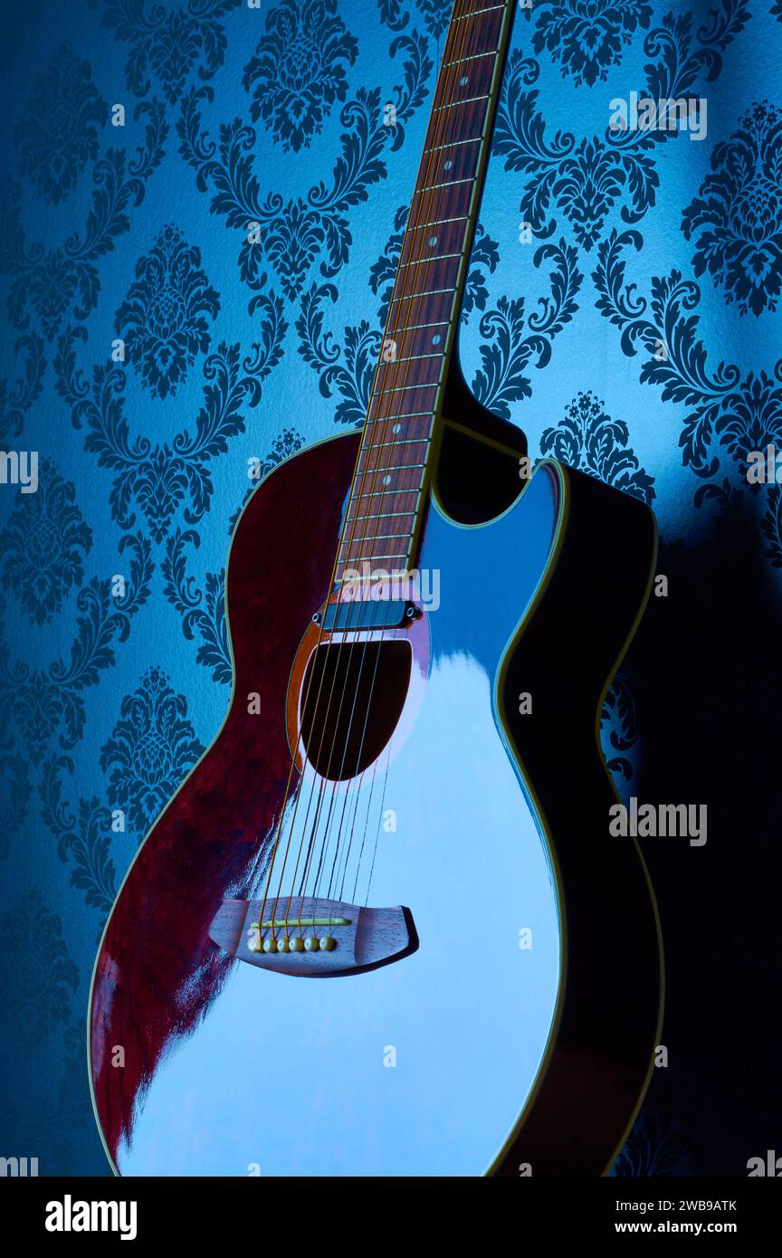 Un'immagine verticale e lunatica di una chitarra elettrica-acustica che riflette la luce blu su uno sfondo blu a motivi di sfondo. Foto Stock