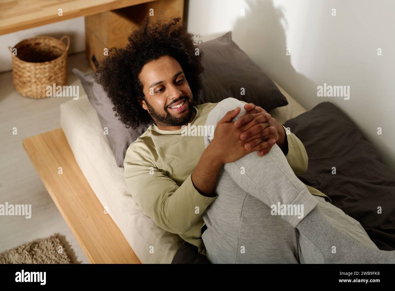 Uomo sorridente in pullover di cotone e pantaloni tuta che fa esercizio di stretching per le gambe dopo il sonno mentre si sdraia a letto la mattina Foto Stock