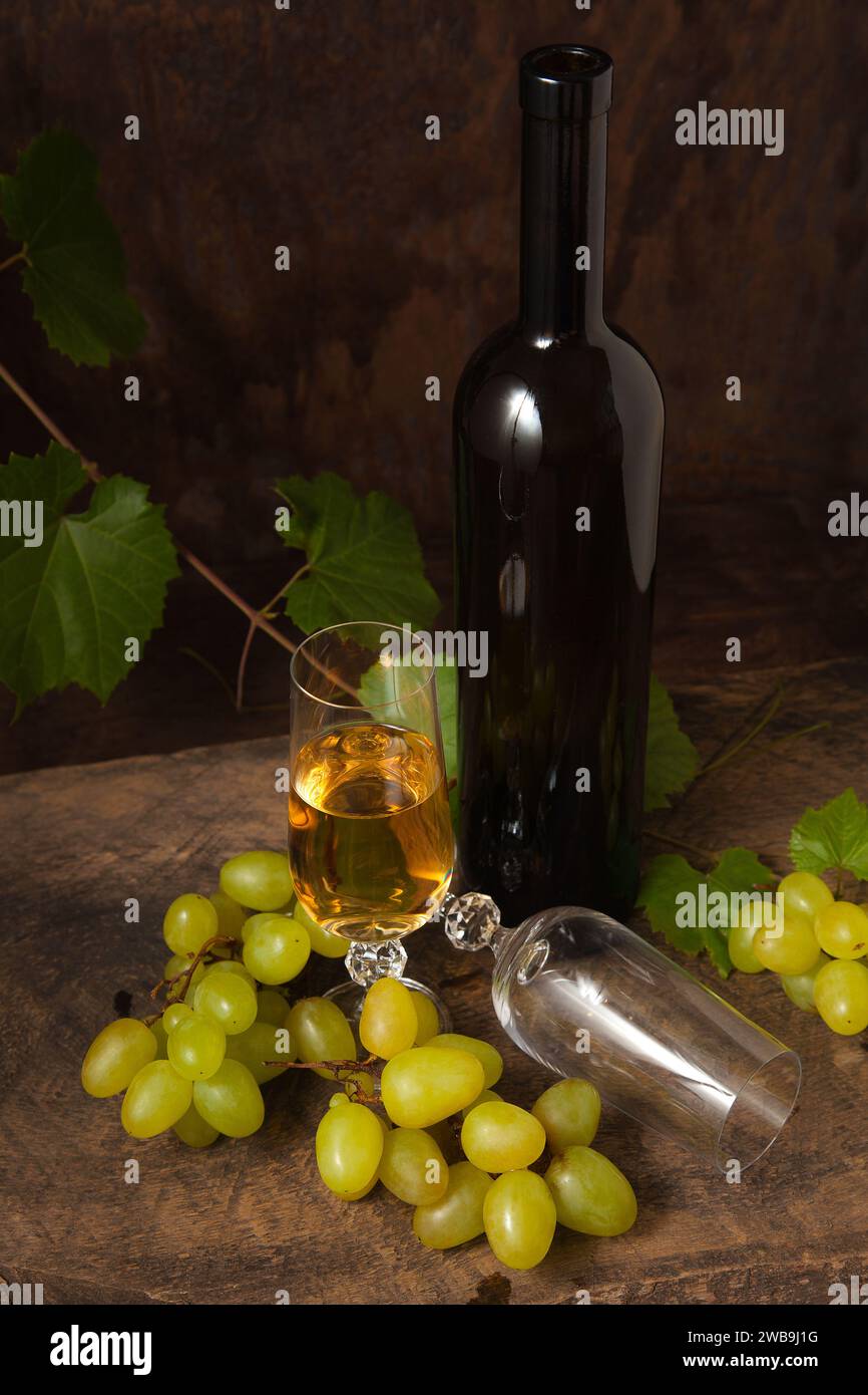 Il concetto di una deliziosa bevanda alcolica, vino e uva. Bottiglia di vino, bicchiere vuoto e bicchiere di vino bianco e mazzo di uva rosa su rustico scuro Foto Stock