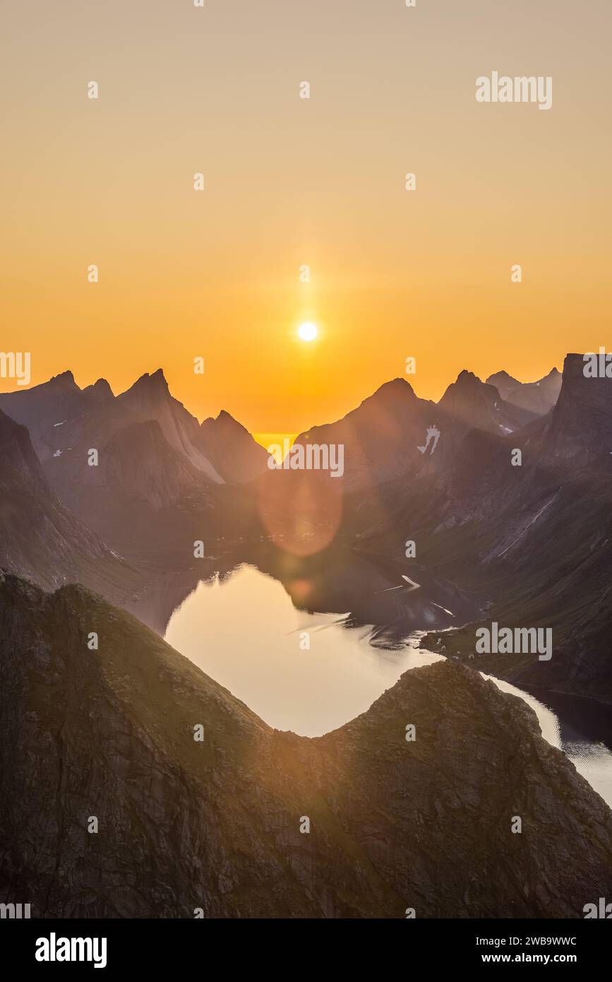 La cornice verticale cattura il sole di mezzanotte emettendo un caldo bagliore sulle picche affilate di Reinebringen nel Lofoten, Norvegia, con il riflesso del sole che brilla Foto Stock