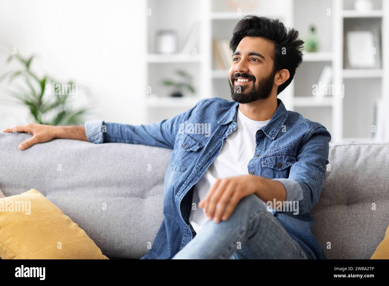 Bellissimo giovane indiano in camicia in denim che si rilassa a casa sul divano grigio Foto Stock