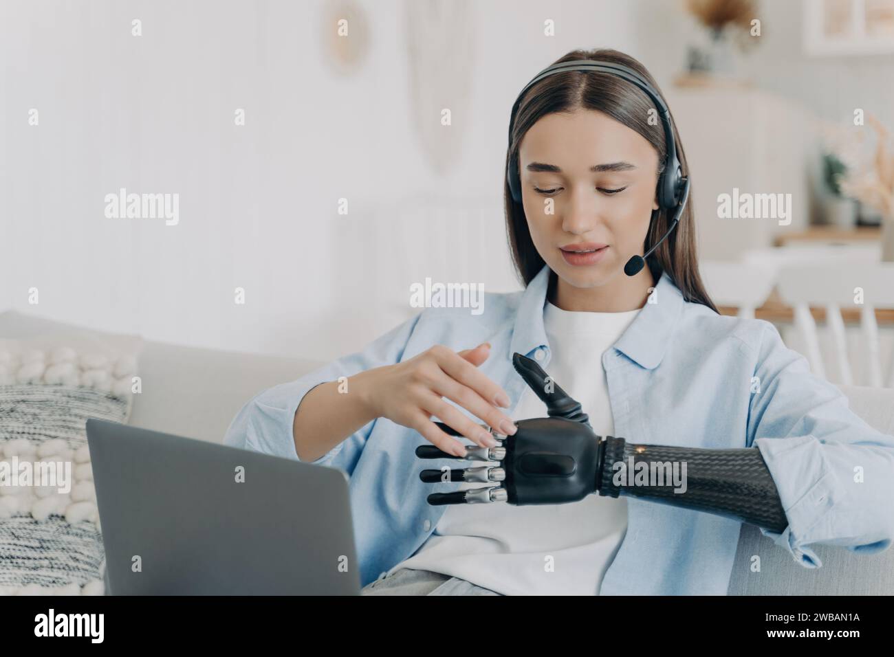 Giovane donna concentrata con braccio bionico che utilizza un computer portatile e cuffie per la comunicazione remota in una casa moderna Foto Stock