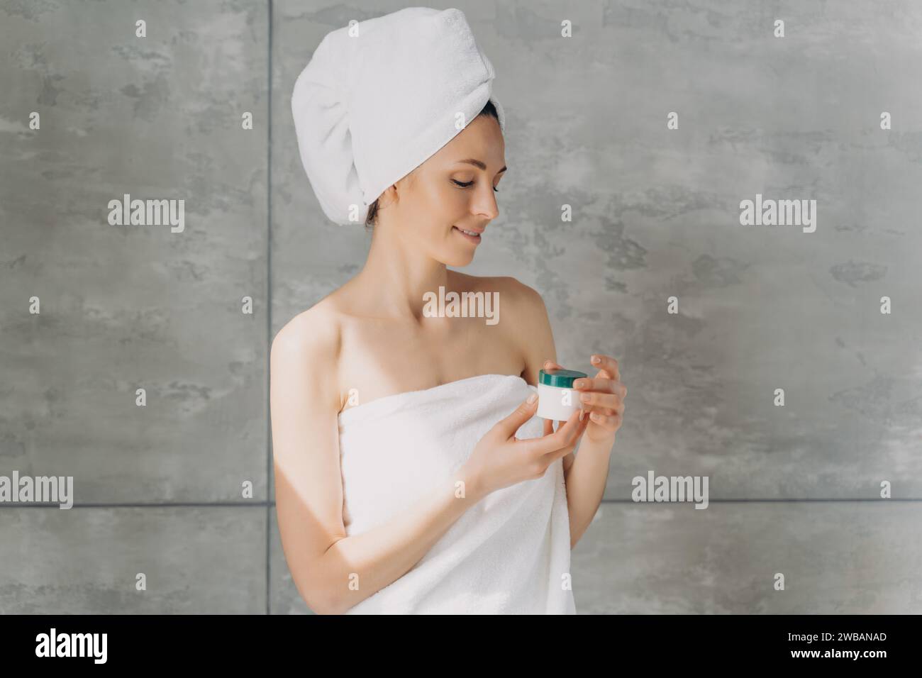 Donna serena avvolta in asciugamani che esaminano i prodotti per la cura della pelle con un sorriso delicato in un ambiente minimalista della spa Foto Stock