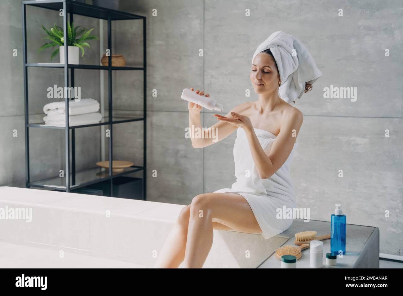 Donna rilassata in un asciugamano che gode di un trattamento di bellezza, tenendo una bottiglia di lozione in un tranquillo bagno in stile spa Foto Stock