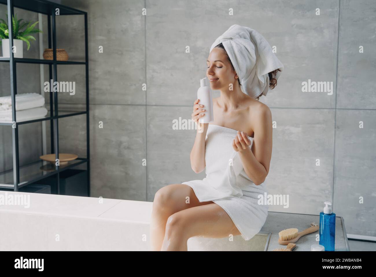 Donna contenta in un centro benessere avvolto in un asciugamano, che apprezza un prodotto per la cura della pelle dall'espressione serena in un ambiente moderno Foto Stock