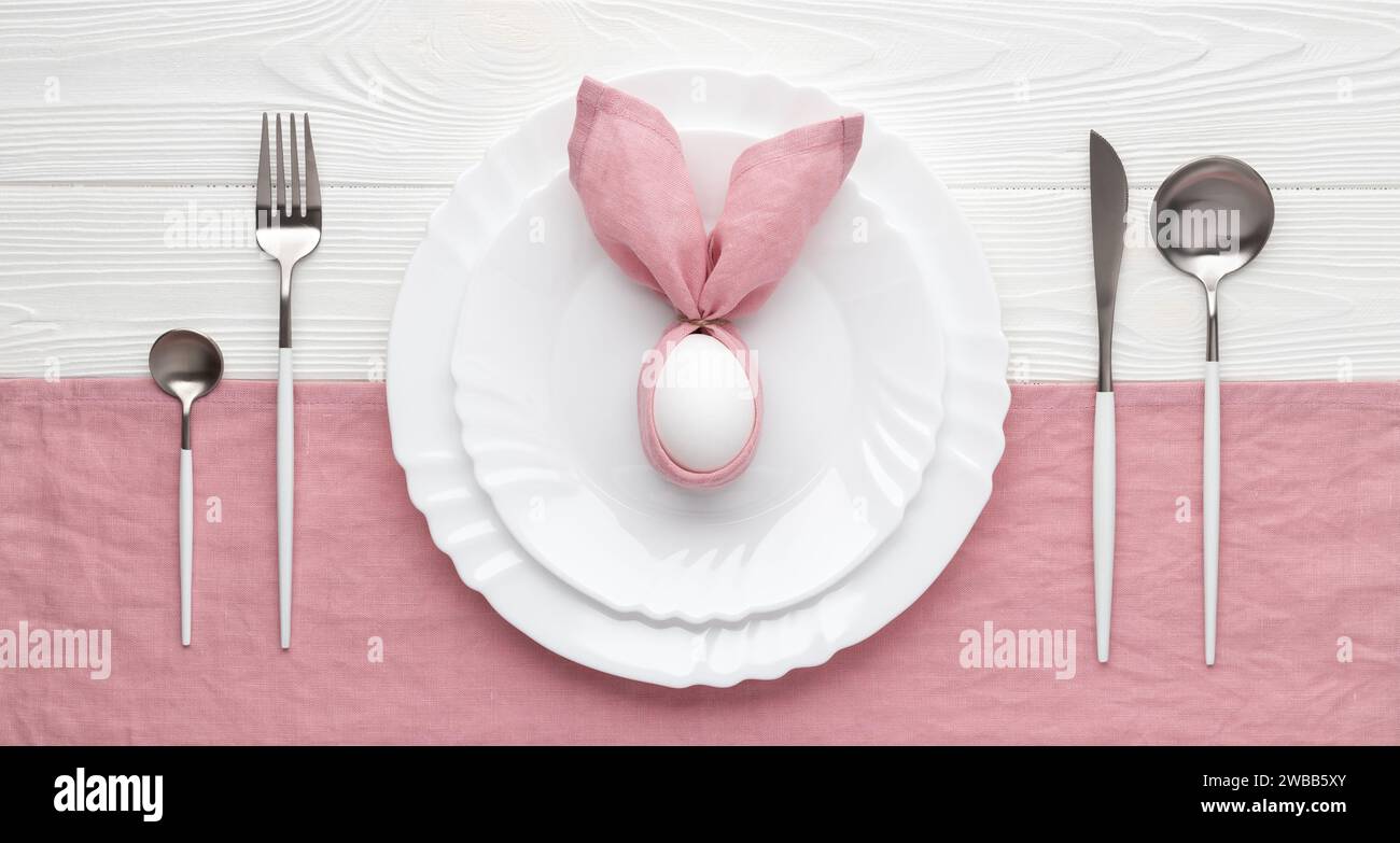Tavolo per le festività pasquali con piatti e posate, concetto di invito. Sfondo di legno bianco. Coniglietto realizzato con tovagliolo e uovo di lino rosa. Vista dall'alto. B Foto Stock
