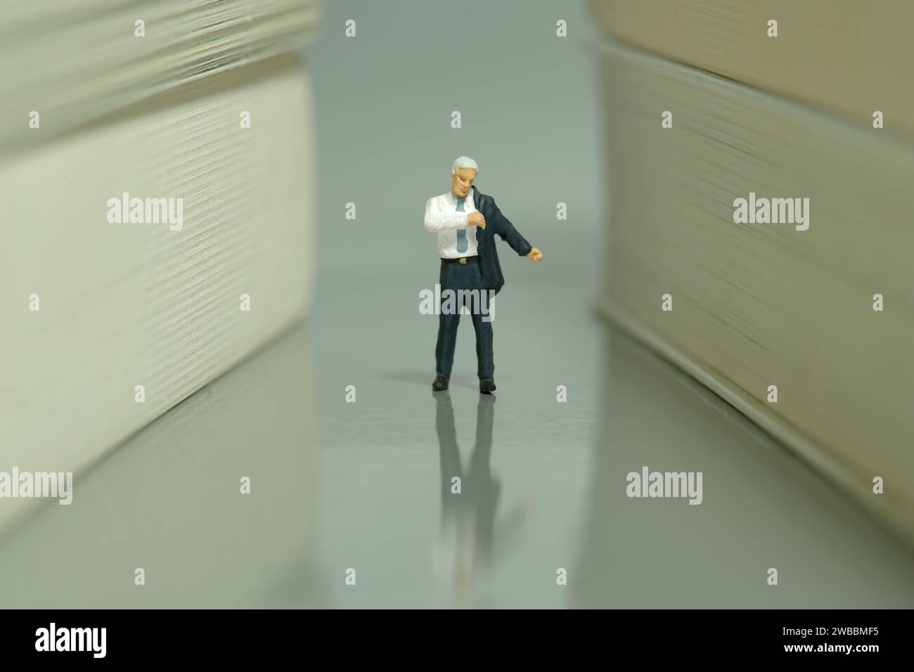 Fotografia di figure giocattolo in miniatura. Un uomo d'affari che si prepara a camminare al corridoio dei libri. Sfondo grigio. Foto immagine Foto Stock