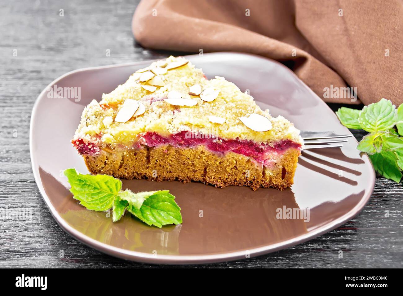 Un pezzo di torta dolce con lamponi, briciole di burro e fiocchi di mandorle in piatto, menta e un asciugamano su fondo di legno Foto Stock