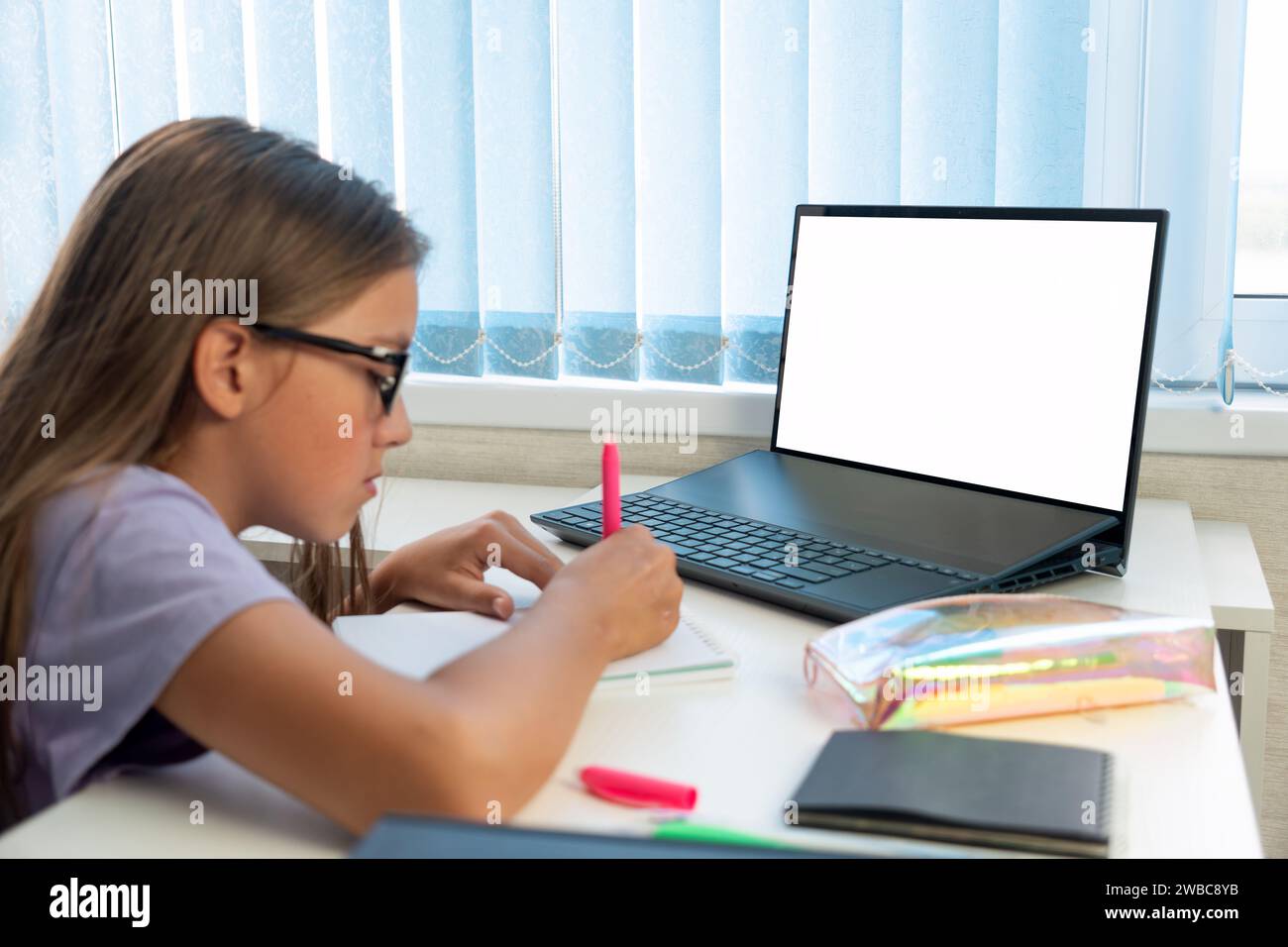 Studente della scuola elementare che scrive in un libro utilizzando un computer portatile. Formazione a distanza online. ragazza che studia a casa con un tablet digitale e. Foto Stock