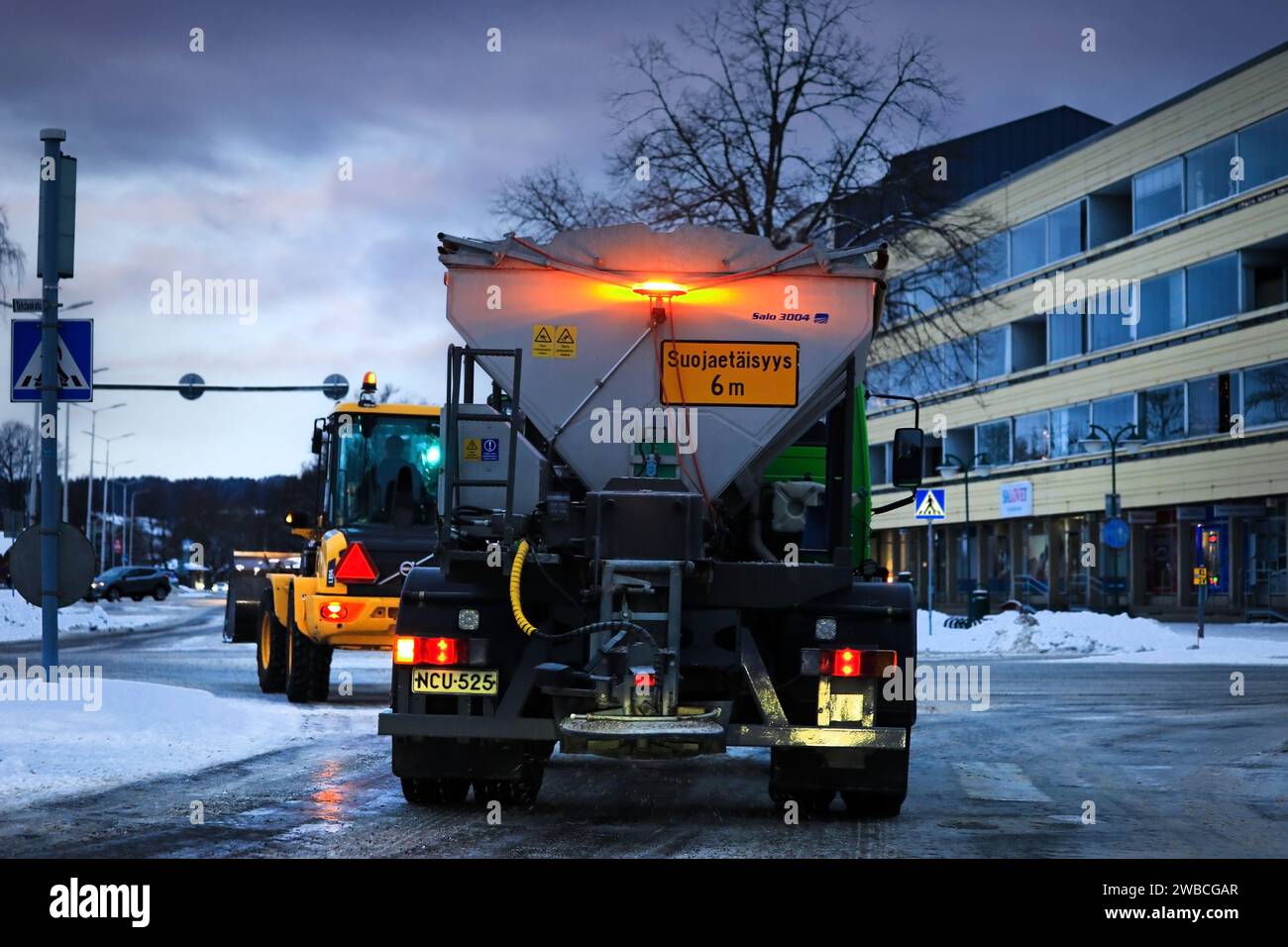 Graffiare il dumper su strada con il trattore da neve davanti, svoltare a sinistra ai semafori. Vista posteriore. Salo, Finlandia. 29 dicembre 2023. Foto Stock