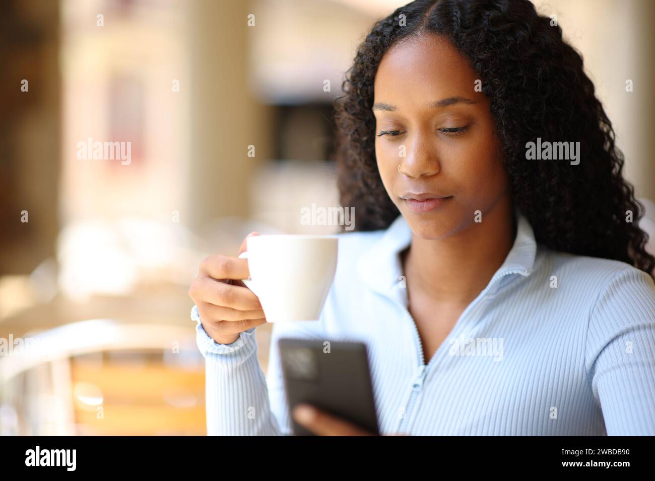 Donna nera che beve un caffè mentre controlla il telefono in una terrazza del ristorante Foto Stock