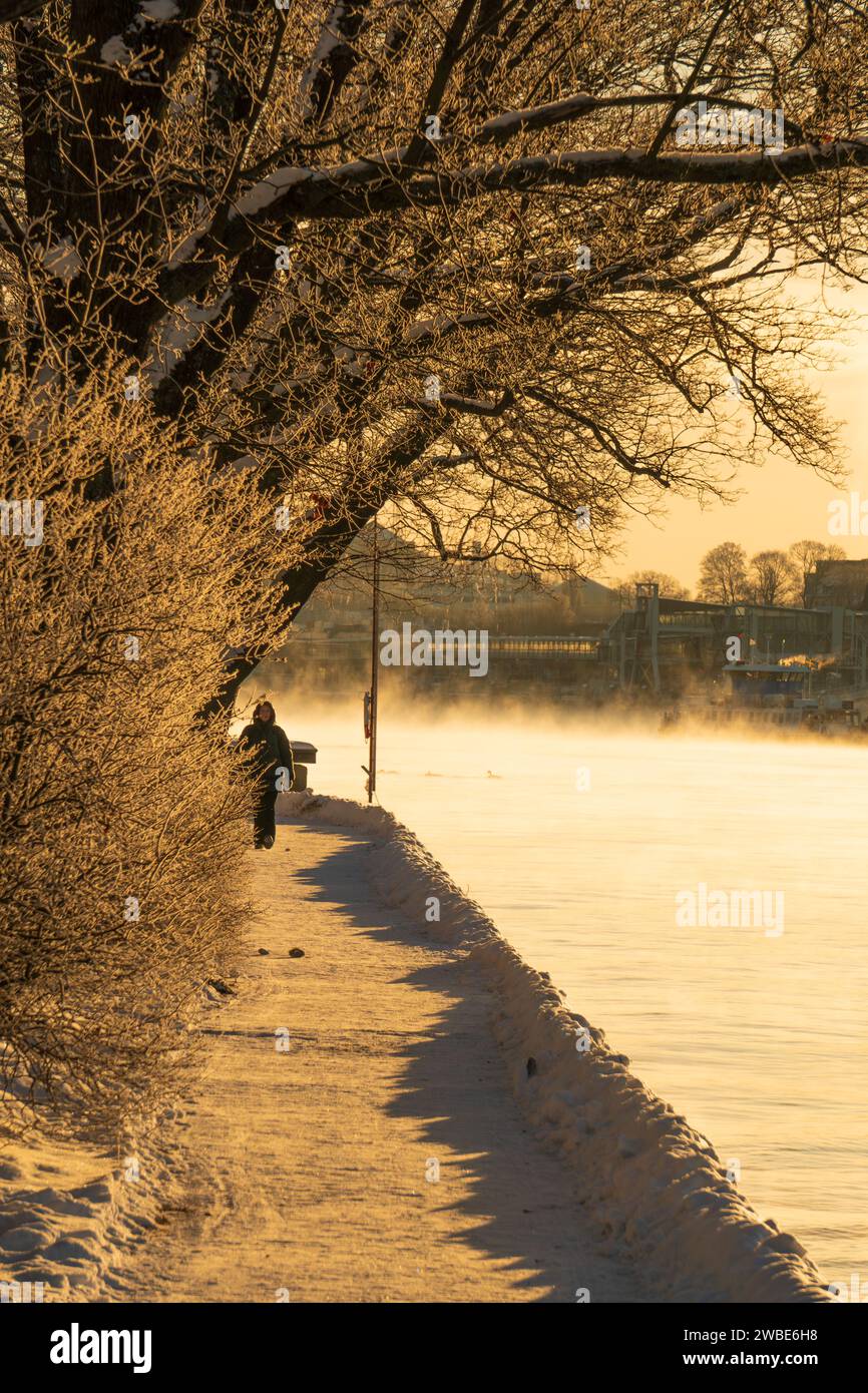Vista di un sentiero in inverno sull'isola di Skeppsholmen, Stoccolma, con fumo di mare visibile dal vento freddo. Una donna che cammina sotto il sole d'inverno. Foto Stock