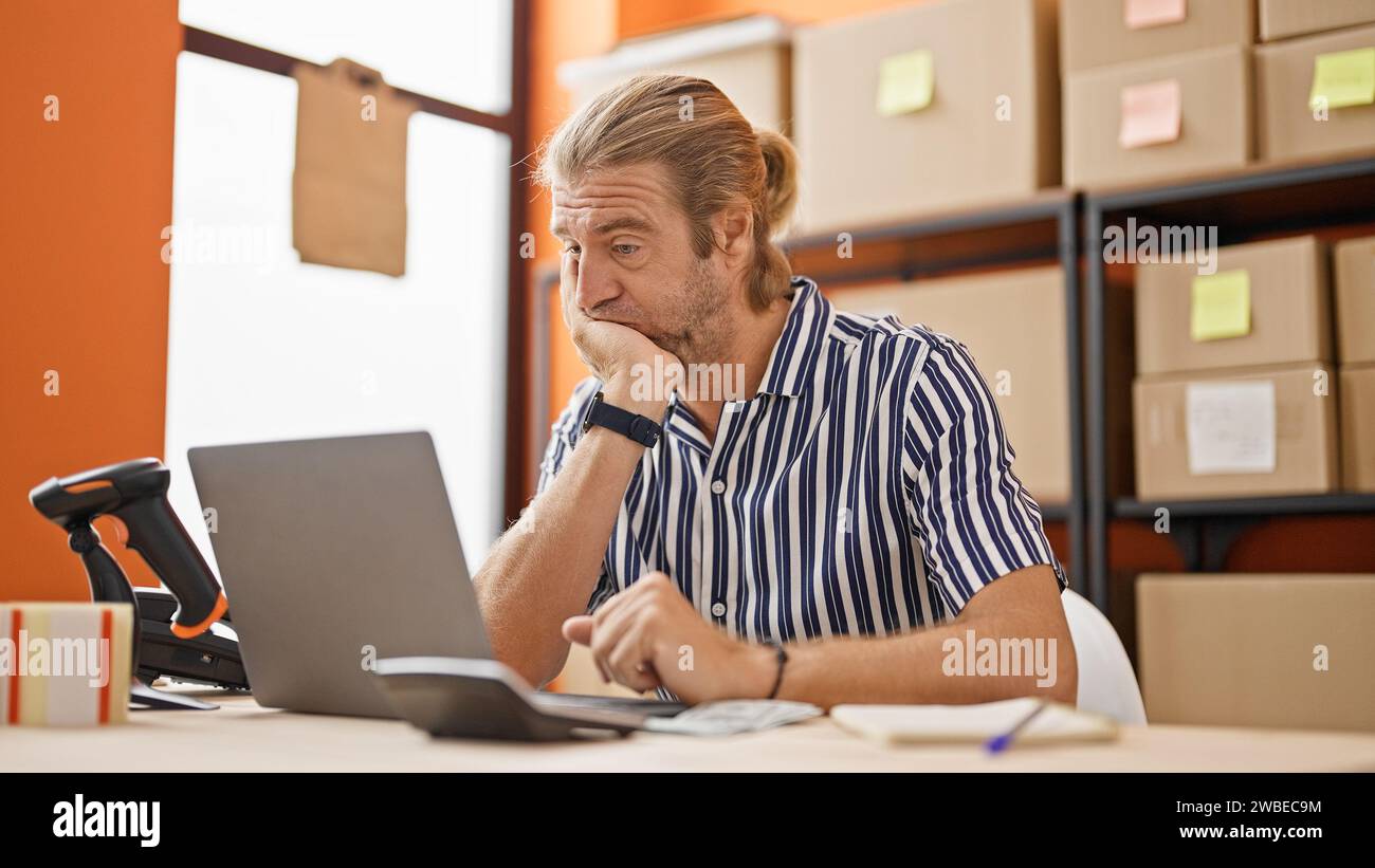 Un uomo preoccupato con capelli lunghi che lavora su un computer portatile in un magazzino circondato da scatole di cartone. Foto Stock