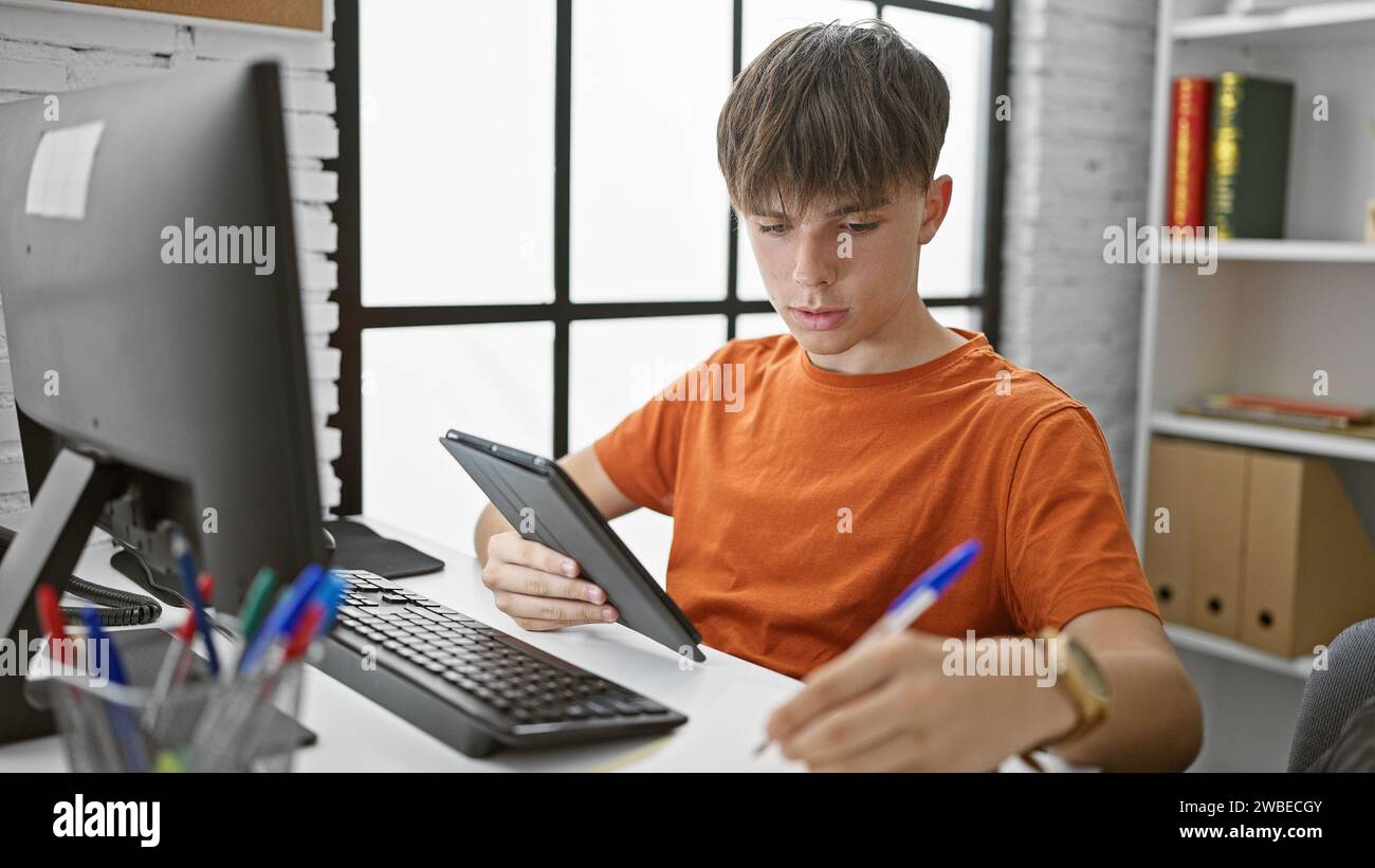 Un giovane studente caucasico studia con un tablet a un tavolo della biblioteca universitaria, concentrato e serio. Foto Stock