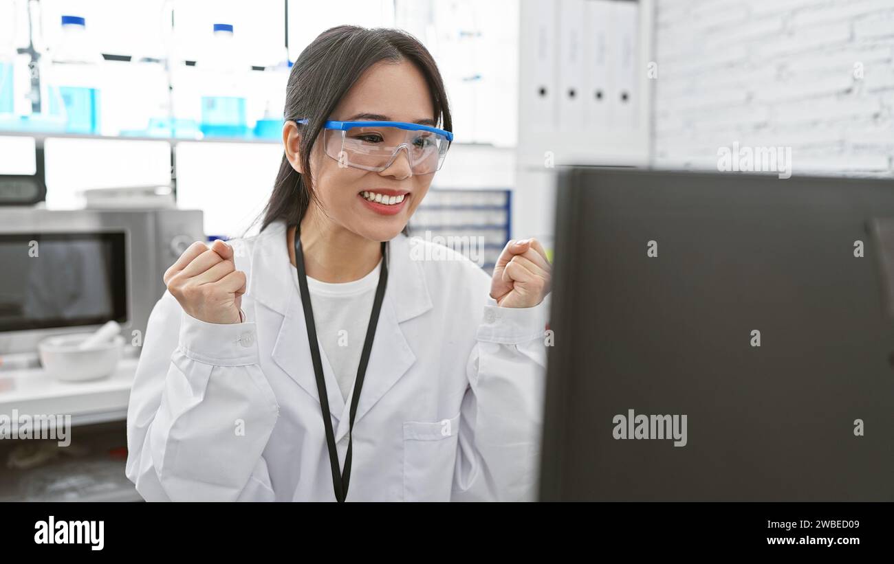 Una scienziata asiatica entusiasta che indossa un camice da laboratorio e occhiali di sicurezza in laboratorio celebra il successo Foto Stock