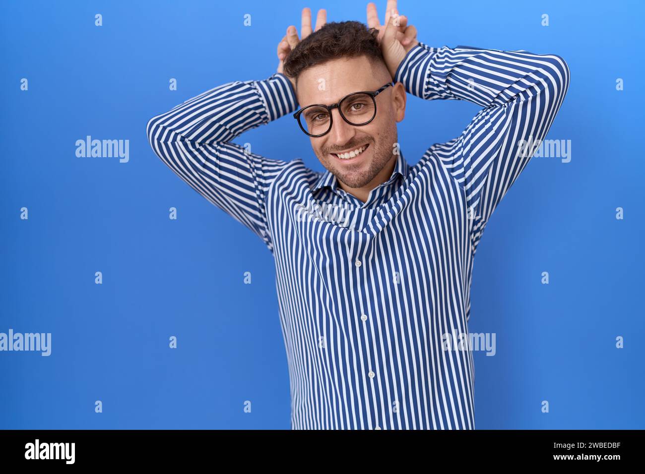 Uomo ispanico con la barba che indossa gli occhiali in posa divertente e pazzo con le dita sulla testa come orecchie coniglietto, sorridente allegro Foto Stock