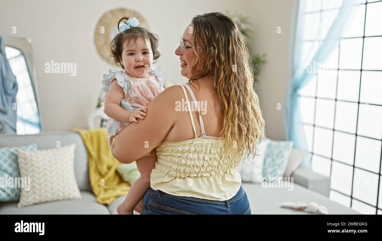 Mamma felice in piedi a casa, tenendo la sua adorabile bambina in braccio, godendo l'amore della famiglia insieme nel loro soggiorno interno con un Foto Stock