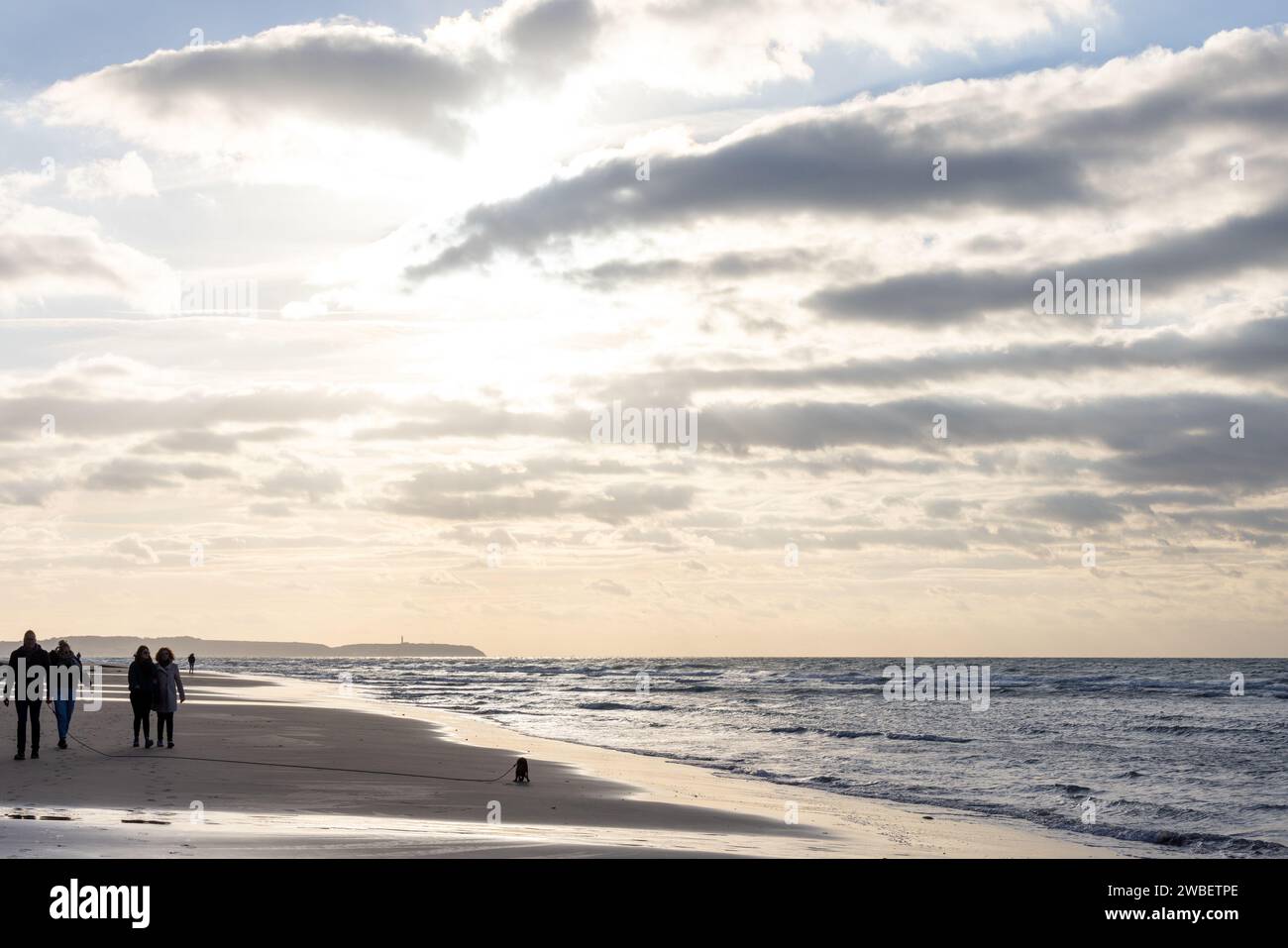 Questa immagine racchiude l'esperienza tranquilla di una passeggiata sulla spiaggia sotto un vasto ed espressivo cielo. Un piccolo gruppo di persone e un cane solitario sulla spiaggia creano un senso di scala e compagnia, sullo sfondo dell'ampio oceano e delle nuvole dinamiche. Il modo in cui la luce del sole filtra attraverso la copertura delle nuvole e illumina la scena aggiunge una qualità paradisiaca alla fotografia, con il faro distante che completa la classica narrazione costiera. Ocean Whispers: Passeggia sulla spiaggia. Foto di alta qualità Foto Stock