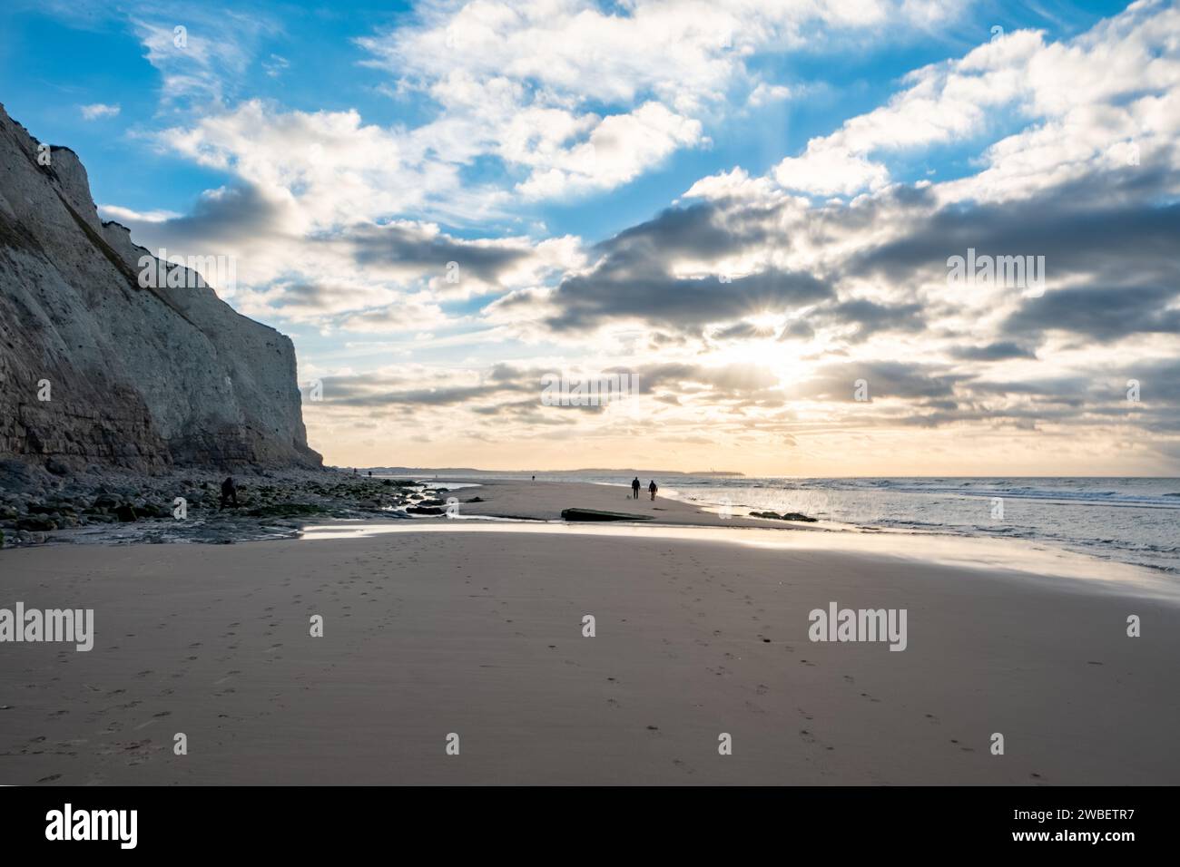 Questa fotografia cattura la grandezza di un paesaggio costiero al tramonto. Due figure distanti sono viste camminare vicino al bordo dell'acqua, fornendo un senso di scala contro l'imponente parete rocciosa. Il sole sbircia attraverso le nuvole sparse, creando un riflettore celeste sull'oceano. Le impronte sulla sabbia suggeriscono la natura transitoria della presenza umana in contrasto con l'erosione senza tempo delle scogliere. Figure lontane e cieli spettacolari. Foto di alta qualità Foto Stock